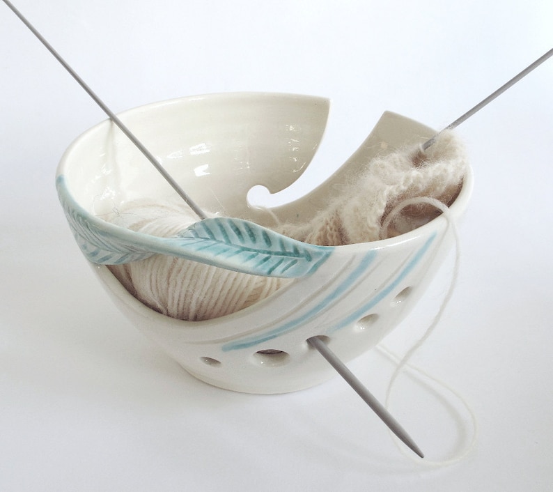 Op de afbeelding: Witte keramische garenkom met een turquoise bladerenontwerp en gaten waar garen doorheen kan worden gevoerd. Twee breinaalden bevinden zich in de kom, een met garen.