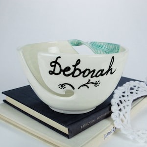 Op de afbeelding: Witte keramische garenkom met een groen bladerenontwerp en de naam "Deborah" in het zwart geschreven. De kom heeft een gat aan de zijkant waar garen doorheen kan.