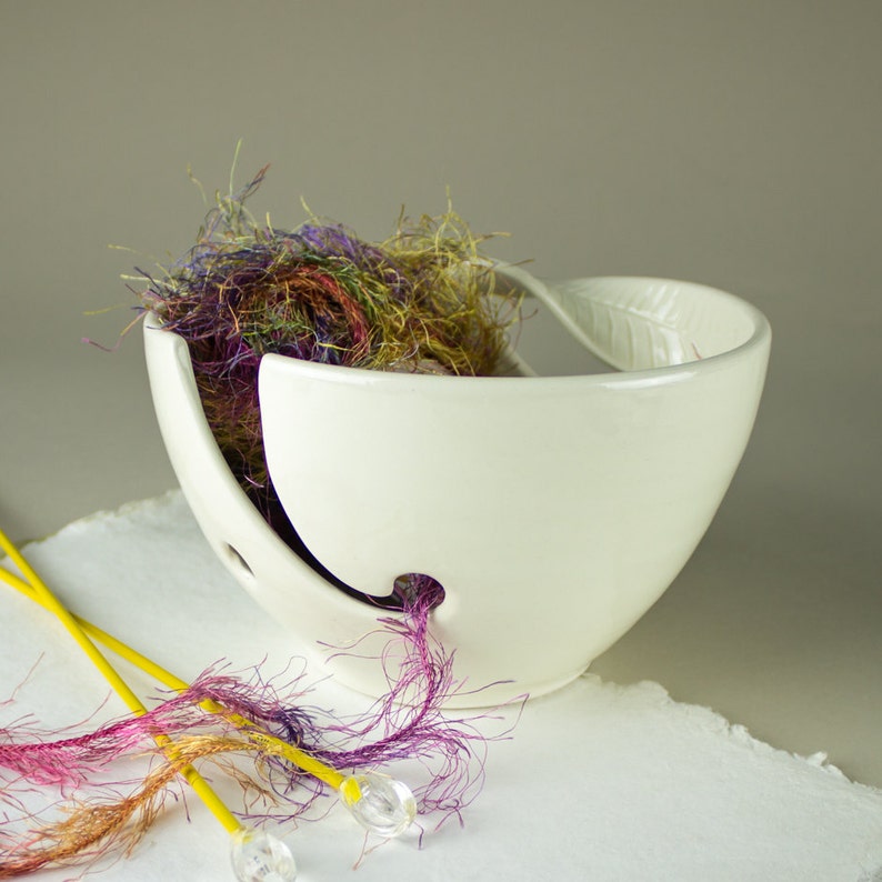May include: A white ceramic yarn bowl with a leaf-shaped handle, filled with colorful yarn. Yellow knitting needles with clear, rounded tips are in the foreground. The bowl has a hole to feed the yarn through.