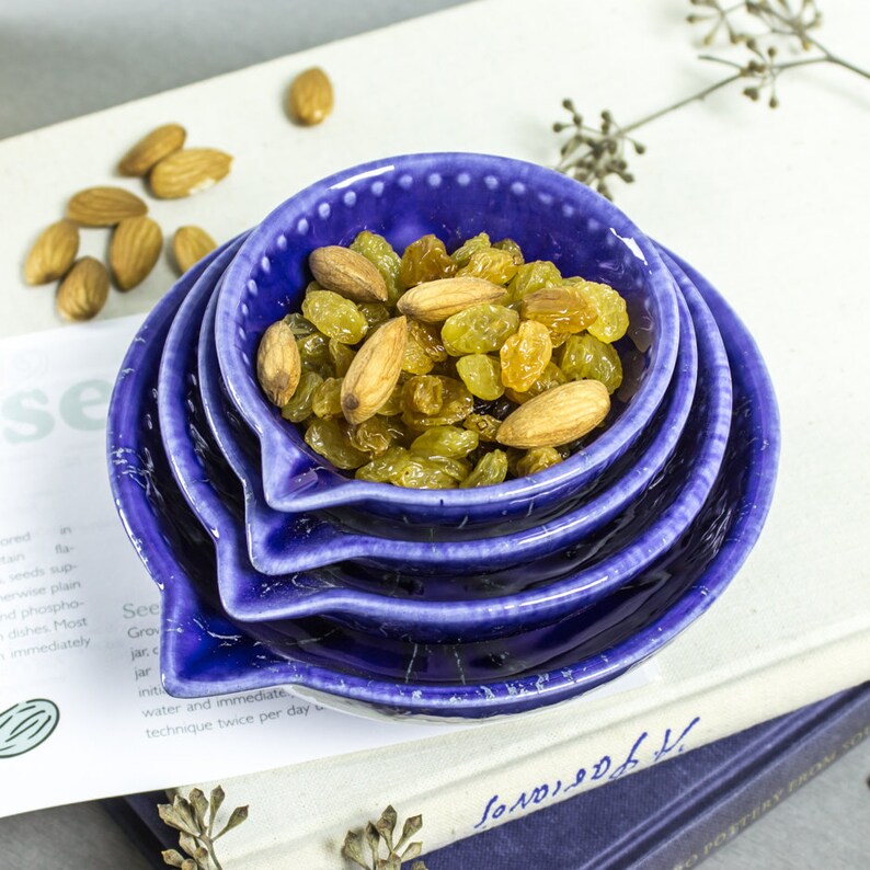 May include: Four blue ceramic nesting bowls with a white speckled glaze. The top bowl is filled with golden raisins and almonds.