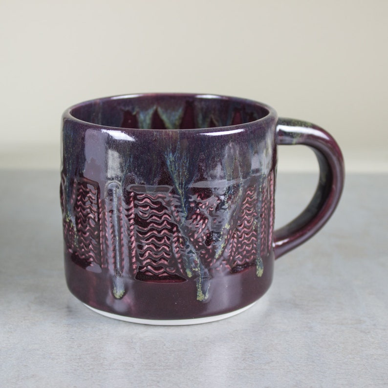 May include: A handcrafted ceramic mug with a deep purple glaze. The mug features a textured design with vertical and wavy patterns. The handle and interior have a glossy finish, and the base is white. A unique piece for enjoying hot beverages.