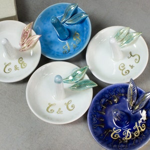 May include: A collection of ceramic ring holders in various colors, including white and blue. Each holder features a central ring post and is adorned with leaf-shaped accents. Gold initials are painted on the surface of each holder.