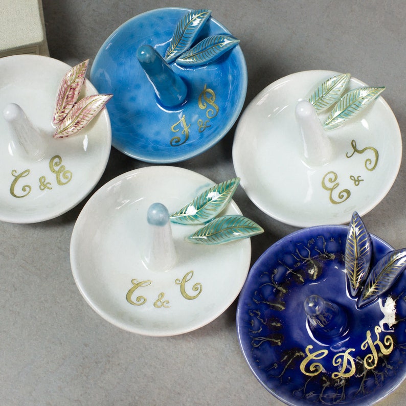 May include: A collection of ceramic ring holders in various colors, including white and blue. Each holder features a central ring post and is adorned with leaf-shaped accents. Gold initials are painted on the surface of each holder.