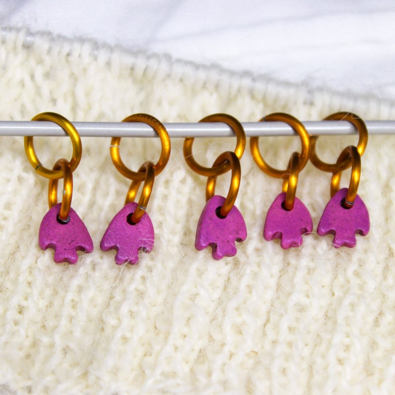May include: Five gold stitch markers with pink heart-shaped beads. The markers are strung on a silver knitting needle.