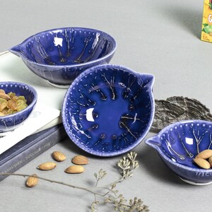 May include: Three blue ceramic bowls with a white crackle glaze design. The bowls are different sizes and have a pour spout. The bowls are on a table with almonds and a book.