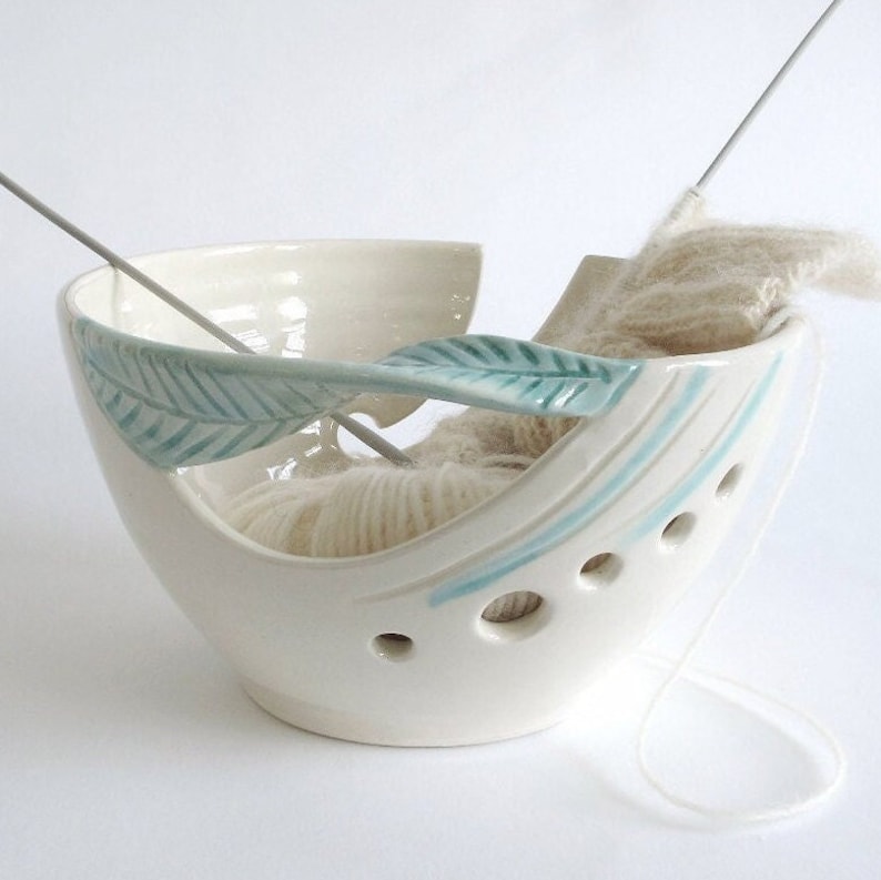 May include: White ceramic yarn bowl with teal leaf design and holes for yarn to pass through. A ball of yarn and knitting needles are in the bowl.