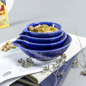 May include: A set of four blue ceramic measuring cups with a speckled glaze. The cups are nested together and filled with a mixture of almonds and golden raisins.