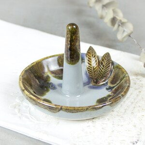 May include: A ceramic ring holder with a light blue glaze and brown and green accents. The holder features a raised ring stand and two leaf-shaped details.