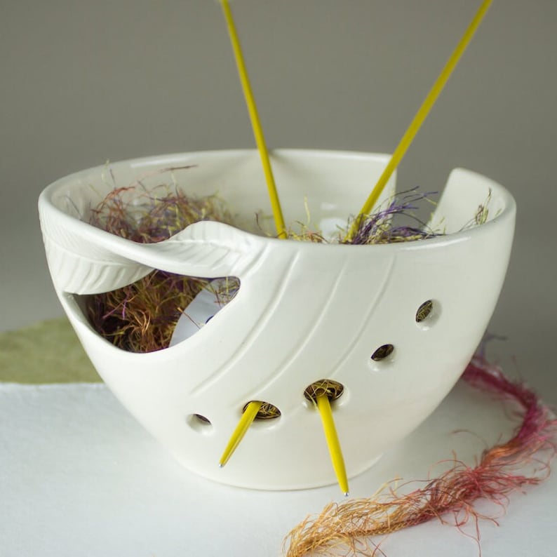 May include: A white ceramic yarn bowl with a leaf design and holes for needles. The bowl contains yellow knitting needles and colorful yarn. The bowl is designed to hold yarn while knitting or crocheting.