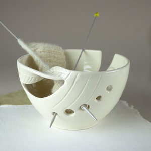 May include: Cream-colored ceramic yarn bowl with a feather-shaped cutout and carved lines. The bowl holds a small amount of off-white yarn and two knitting needles. The bowl has three small holes for the needles. A craft item.