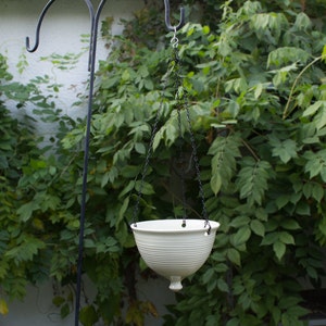 May include: A white ceramic hanging planter with a ribbed design hangs from a black metal chain against a backdrop of green foliage.