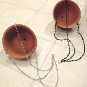 May include: Two terracotta hanging planters with silver and black chains. The planters are empty and ready to be filled with plants.
