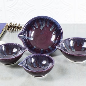 Op de afbeelding: Een set van vier keramische maatbekers in verschillende maten. De kommen zijn diep paars met een glanzende afwerking en witte en blauwe accenten. De kleinste kom is gemarkeerd met "1/4 CUP".