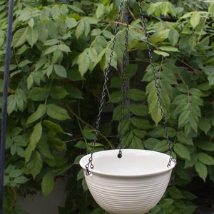 May include: White ceramic hanging planter with a ribbed design and a small knob at the bottom. The planter is suspended from a black chain.