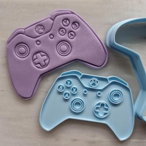Może przedstawiać: Dwa plastikowe wykrawacze do ciasteczek w kształcie kontrolera do gier wideo. Górny wykrawacz jest fioletowy, a dolny jasnoniebieski.