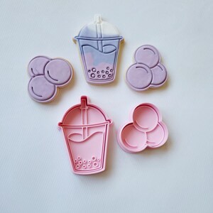 May include: Five cookie cutters in the shape of bubble tea and boba. Two cutters are pink, and three are purple. The bubble tea cutters have a cup shape with a straw. The boba cutters have a three-circle design.