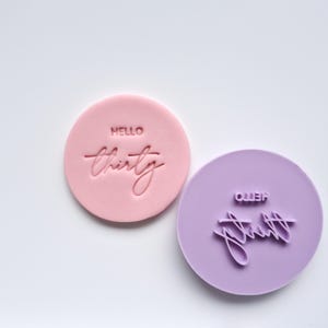 May include: Two circular cookie stamps, one pink and one lilac. The pink stamp reads "HELLO thirty" in a script font. The lilac stamp also reads "HELLO thirty" in a script font. The stamps are on a white background.