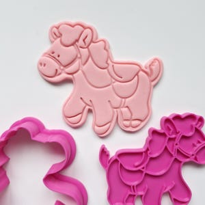 May include: Three pink plastic horse-shaped cookie cutters. The largest cutter is a detailed horse with a saddle. The other two cutters are a lighter pink and a darker pink, and are also horse-shaped. The cutters are on a white background.