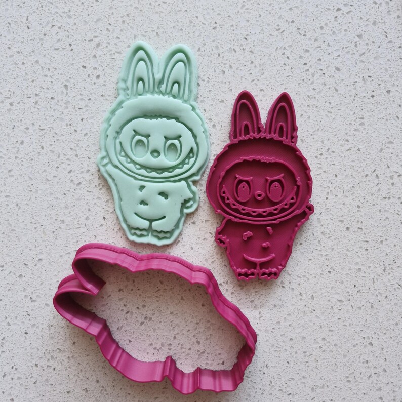 May include: Three cookie cutters in the shape of a cartoon bunny character. One is a light green color, another is a deep pink, and the third is a pink outline of the bunny. The bunny has a smiling face and a hooded outfit.