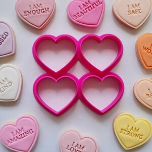 May include: An assortment of heart-shaped biscuits and a pink heart-shaped cookie cutter. The biscuits are in pastel shades, each with an embossed positive message like "I AM ENOUGH" and "I AM BEAUTIFUL". The cookie cutter is a vibrant pink.