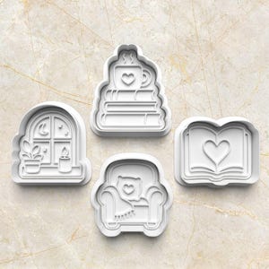 May include: A set of four white plastic cookie cutters featuring designs of a mug on a stack of books, an arched window with a plant and candle, an armchair with a pillow and throw blanket, and an open book with a heart shape in the centre.