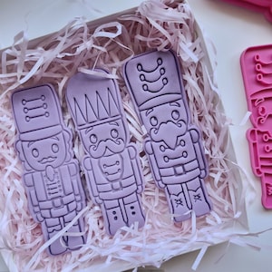 May include: Three lavender-coloured plastic cookie cutters shaped like nutcrackers, presented in a white box with pink paper shreds. The cutters feature detailed designs, including hats, moustaches, and uniforms. A pink cutter is visible on the right.
