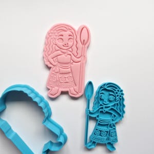May include: Pink and blue plastic cookie cutters in the shape of a cartoon character holding a spear. The pink cutter is a solid form, while the blue cutter is an outline. The cutters are on a white surface.
