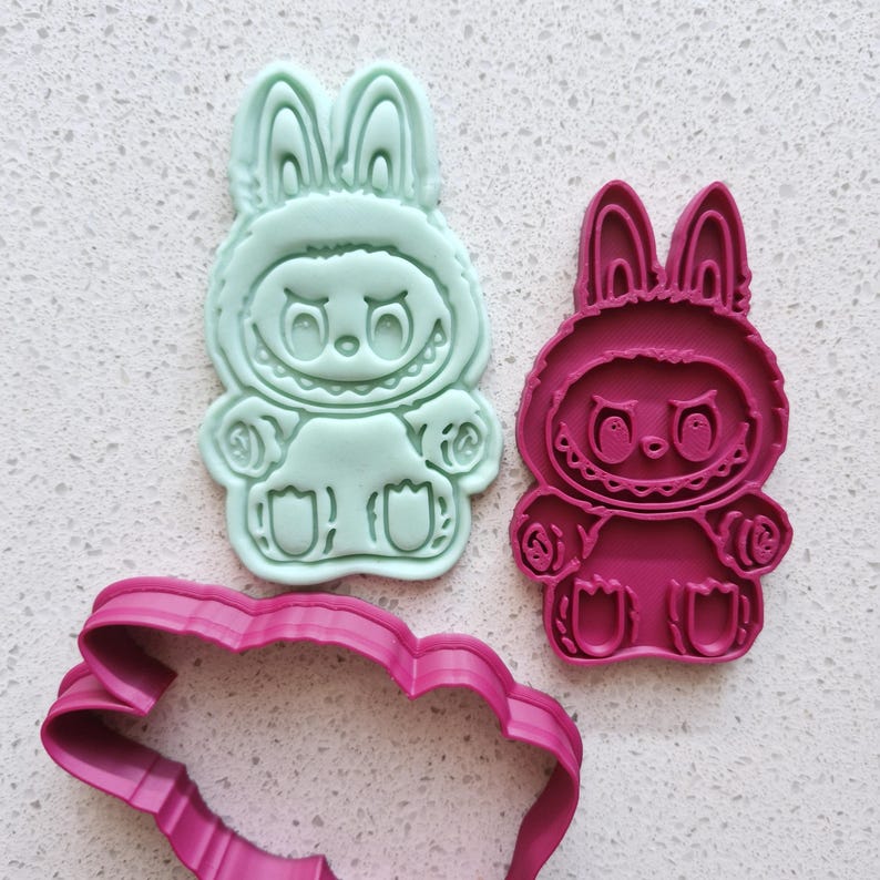 May include: Two bunny-shaped cookie cutters in mint green and magenta, alongside a magenta scalloped edge cutter. The bunny design features a hooded figure with a smiling face and rounded paws. Ideal for baking themed treats.
