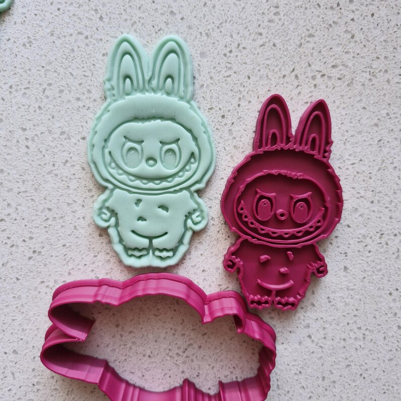 May include: Three cookie cutters in the shape of a cartoon bunny character. One is mint green, one is magenta, and the third is a magenta cloud shape. The bunny has a smiling face and a hood.