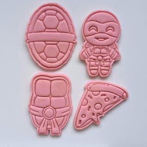 May include: Four pink cookie cutters: a turtle shell, a ninja turtle figure, another shell, and a pizza slice. The cutters are plastic and sit on a white surface. Ideal for making themed biscuits.