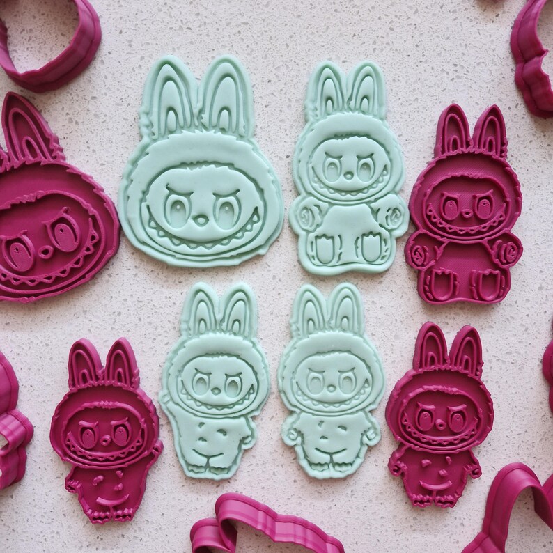 May include: A collection of bunny-shaped cookie cutters in shades of teal and magenta. The cutters feature a cartoon bunny design with a smiling face and long ears. Some cutters are complete bunny figures, while others are just the bunny face.