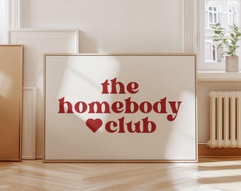 May include: A white framed print with the text "the homebody club" in red lettering with a red heart symbol between the words "homebody" and "club".