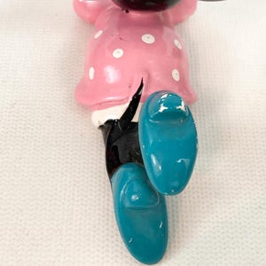 May include: A vintage Minnie Mouse figurine, featuring a pink dress with white polka dots and turquoise shoes. The black legs and white gloves are also visible. The figurine is a classic Disney character.