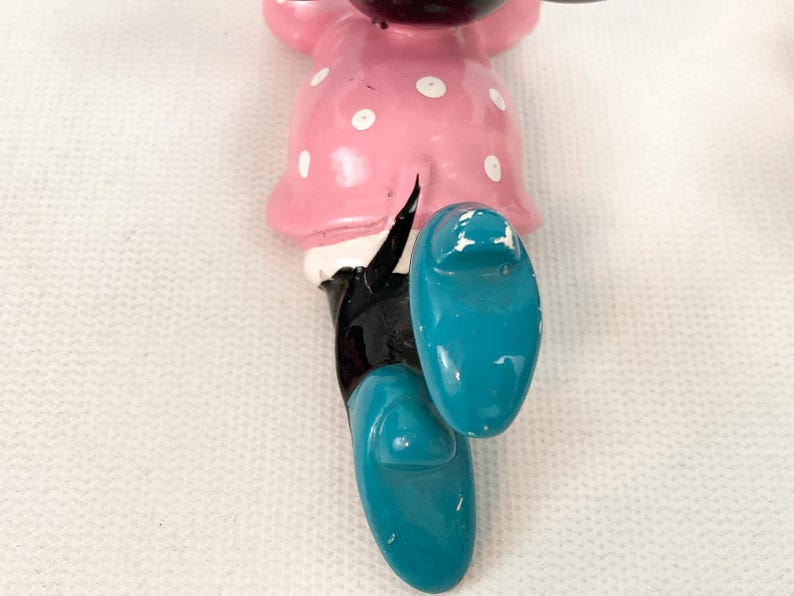 May include: A vintage Minnie Mouse figurine, featuring a pink dress with white polka dots and turquoise shoes. The black legs and white gloves are also visible. The figurine is a classic Disney character.