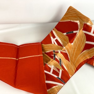 May include: A long, orange fabric sash with a pattern of gold leaves and black stems. The sash is folded in half and the ends are overlapping.