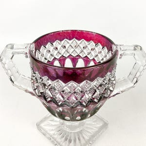 May include: A clear glass sugar bowl with a square base and two handles. The bowl is decorated with a diamond pattern and has a deep red hue.