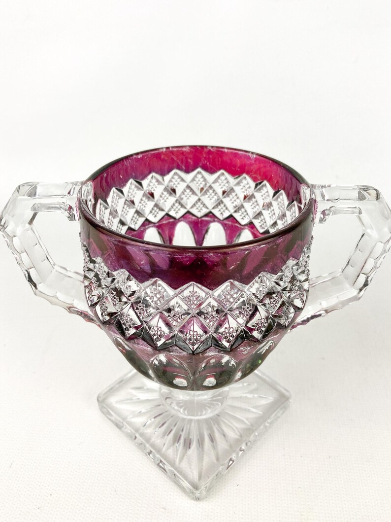 May include: A clear glass sugar bowl with a square base and two handles. The bowl is decorated with a diamond pattern and has a deep red hue.