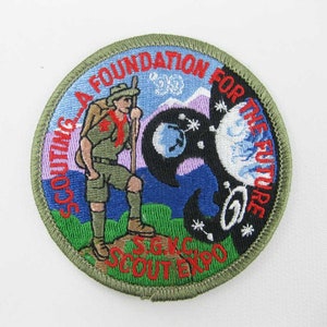 May include: A green, round embroidered patch with the text "SCOUTING A FOUNDATION FOR THE FUTURE" in red and white. The patch features a cartoon scout with a backpack and a staff, standing in front of a blue and purple mountain range. The text "SGKC SCOUT EXPO" is also embroidered on the patch.