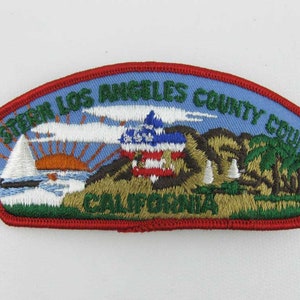May include: Embroidered patch with a blue background and red border. The patch features text that reads "WESTERN LOS ANGELES COUNTY COUNCIL CALIFORNIA". The design includes a sailboat, sunset, and a depiction of the American flag.