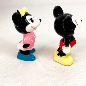 Może przedstawiać: Dwie ceramiczne figurki Myszki Miki i Myszki Minnie. Minnie ma na sobie r&oacute;żową sukienkę w białe grochy, ż&oacute;łtą kokardę i niebieskie buty. Miki ma czerwone szorty i ż&oacute;łte buty. Obie postacie mają czarne uszy i białe twarze.