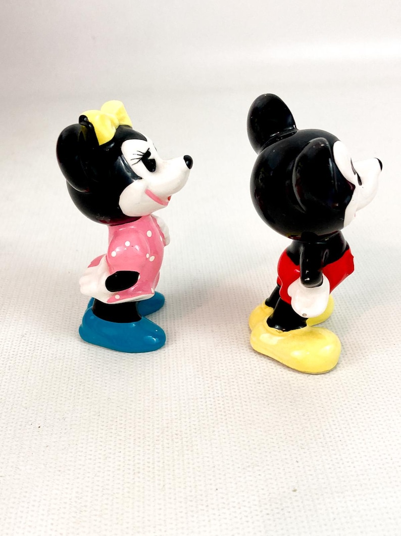 Może przedstawiać: Dwie ceramiczne figurki Myszki Miki i Myszki Minnie. Minnie ma na sobie r&oacute;żową sukienkę w białe grochy, ż&oacute;łtą kokardę i niebieskie buty. Miki ma czerwone szorty i ż&oacute;łte buty. Obie postacie mają czarne uszy i białe twarze.