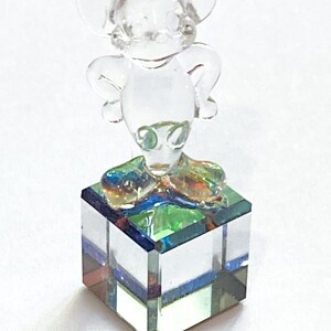May include: A clear glass figurine of a mouse with large ears, standing atop a square, multi-colored glass base. The mouse figure is transparent, and the base features shades of blue, green, and red.