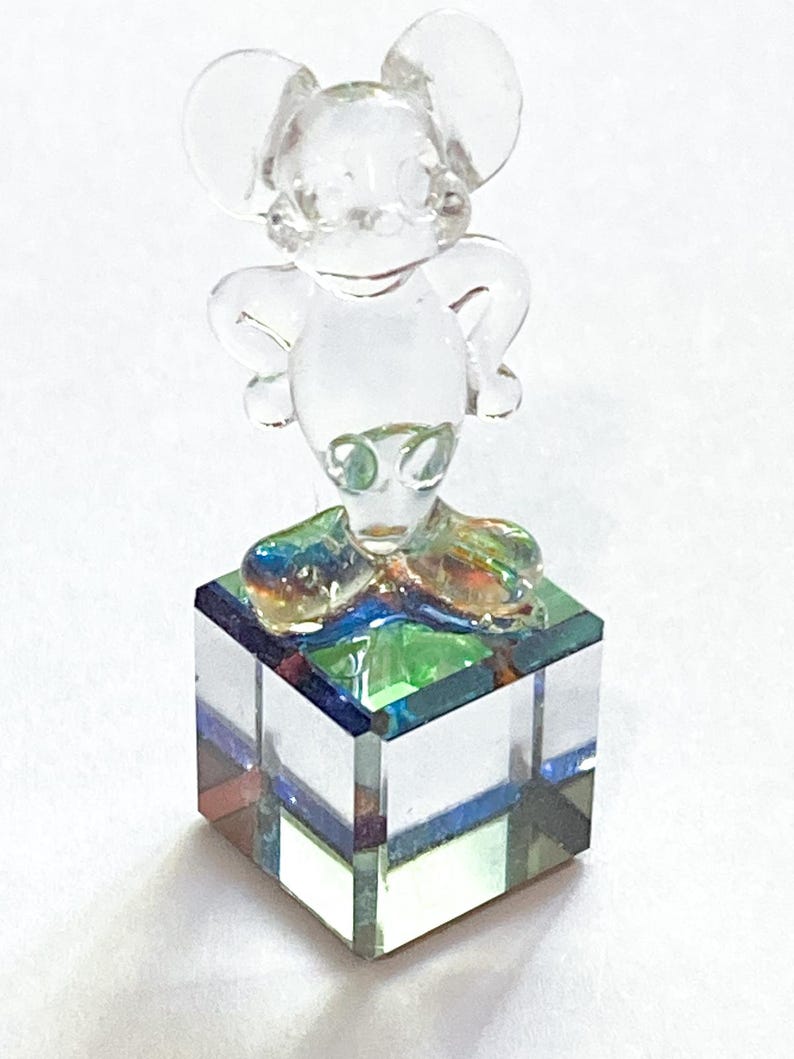 May include: A clear glass figurine of a mouse with large ears, standing atop a square, multi-colored glass base. The mouse figure is transparent, and the base features shades of blue, green, and red.