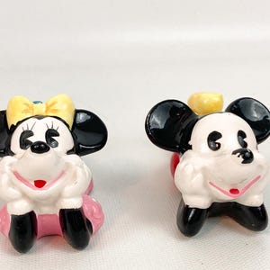 May include: Two ceramic figurines of cartoon characters. One is Minnie Mouse, with a yellow bow and pink skirt. The other is Mickey Mouse. Both have black ears, white faces, and black shoes. They are in a sitting position.