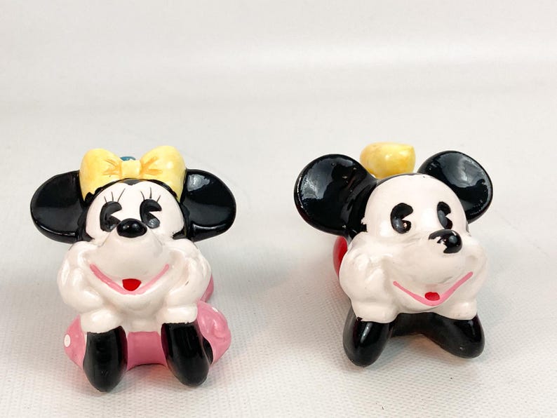 May include: Two ceramic figurines of cartoon characters. One is Minnie Mouse, with a yellow bow and pink skirt. The other is Mickey Mouse. Both have black ears, white faces, and black shoes. They are in a sitting position.