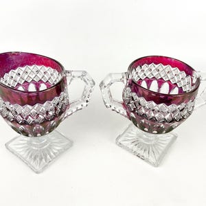 May include: A set of two clear glass creamer and sugar bowls with a footed base. The bowls are decorated with a diamond pattern and have a deep burgundy red color.