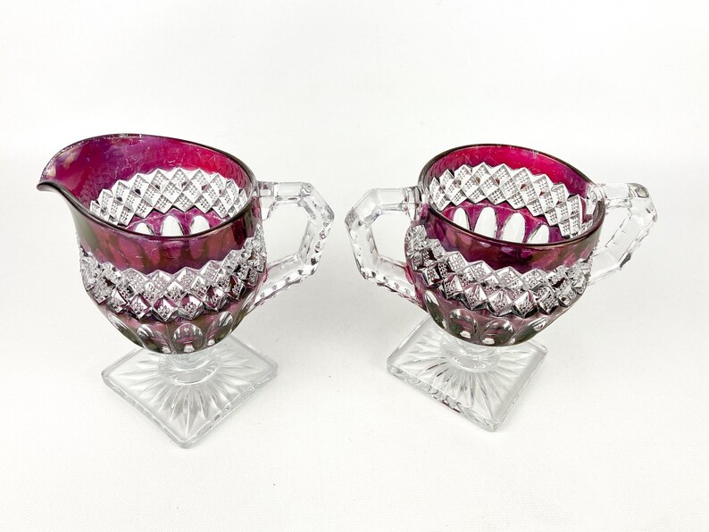 May include: A set of two clear glass creamer and sugar bowls with a footed base. The bowls are decorated with a diamond pattern and have a deep burgundy red color.