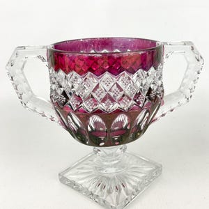 May include: A clear glass pedestal bowl with two handles and a square base. The bowl is decorated with a diamond pattern and has a deep red color.