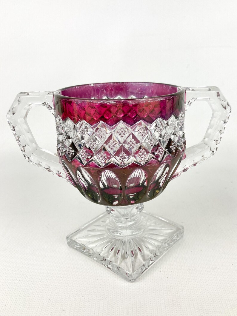 May include: A clear glass pedestal bowl with two handles and a square base. The bowl is decorated with a diamond pattern and has a deep red color.