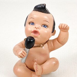 May include: A vintage figurine of a baby with a black mohawk hairstyle, blue eyes, and a gold chain necklace. The baby is holding a black microphone with a silver handle and has one arm raised.